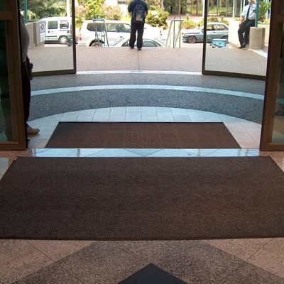 שני שטיחי סף כהים מונחים בכניסה לבניין עם דלתות זכוכית. בחוץ, ברחוב, נראים כמה אנשים ומכוניות.